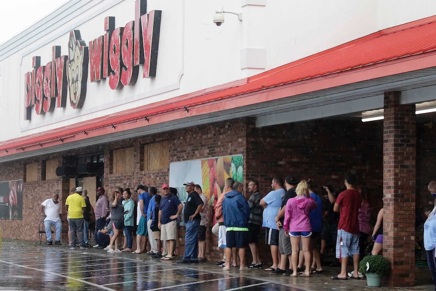 A long line of people stretches out of frame, as people wait for the Piggly Wiggly grocery store to open. Some wear raincoats.