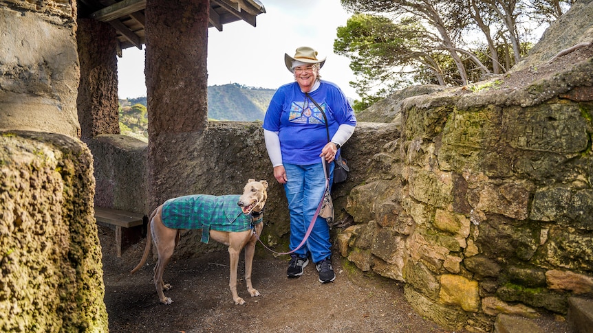 A woman wearing a blue shirt and wide brown hat stands by a mossy stone wall on a sunny day smiling.