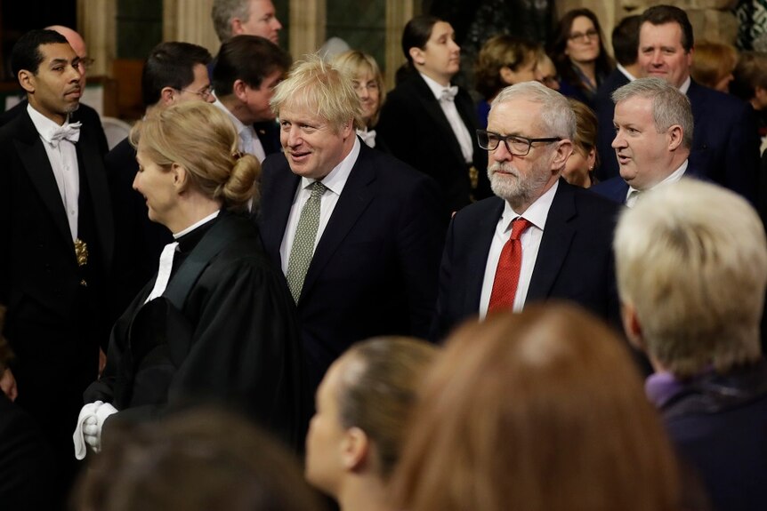 Boris Johnson, smiling, and Jeremy Corbyn, frowning, walk among a crowd.