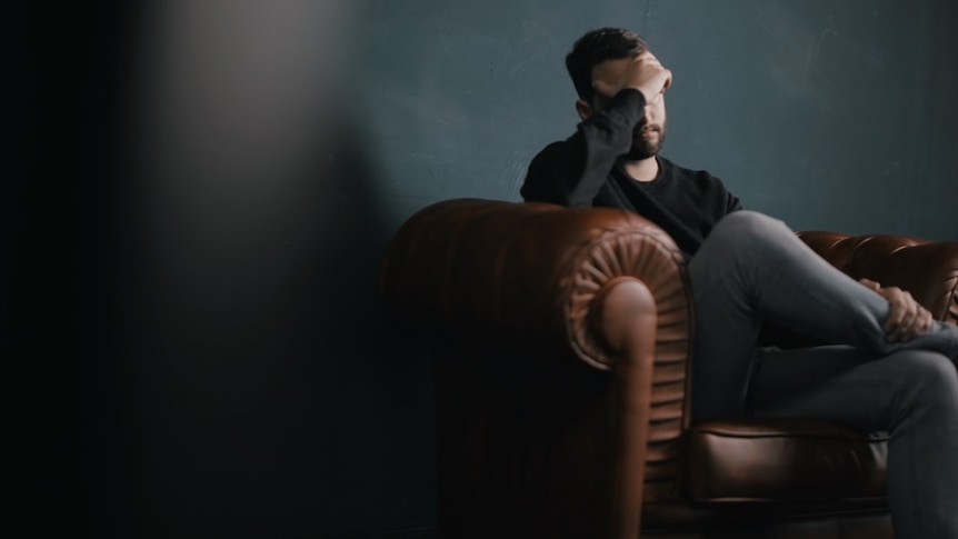 A man sits on a sofa holding his head in his hands.