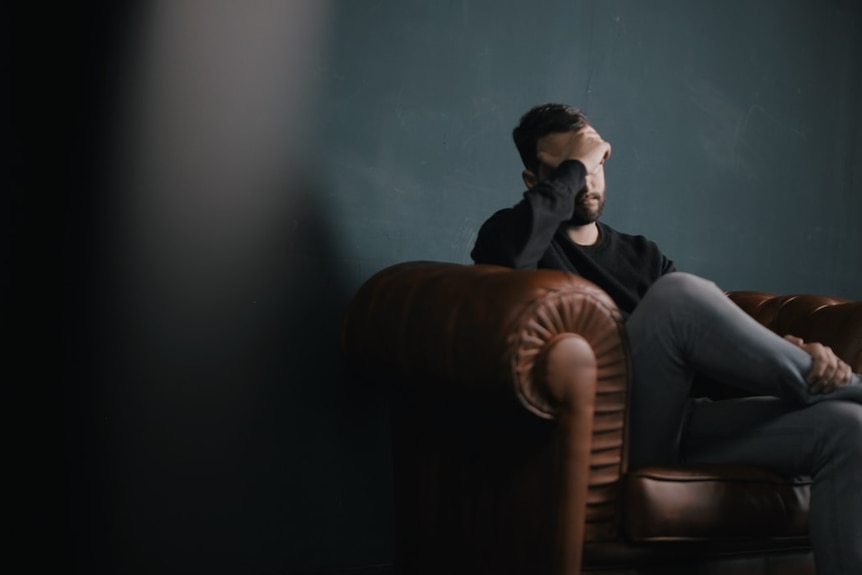 A man sits on a sofa holding his head in his hands.