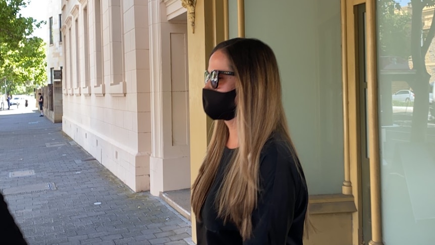 A woman wearing a black shirt, a black mask and sunglasses walks out of a window