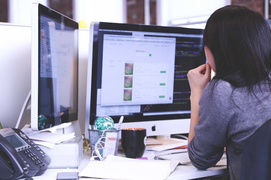 A woman looks at a computer screen in an office setting. A landline phone and mobile phone are on her desk.