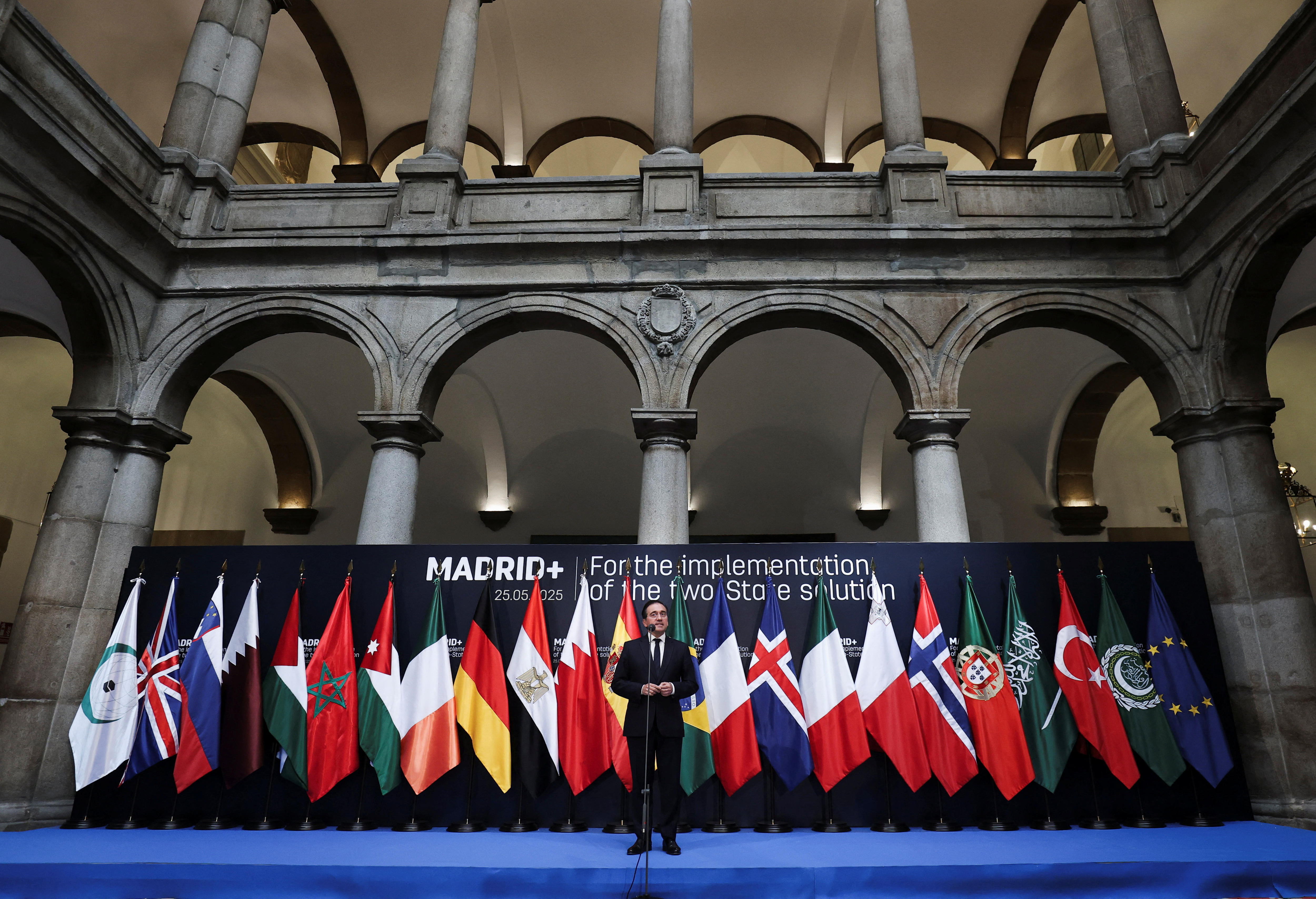 A man in a suit on a blue stage at a microphone in front of a line of European and Arab flags and large concrete archways