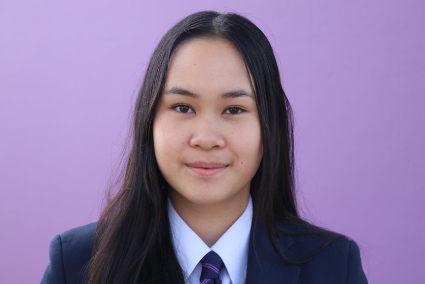A close up of Kathy wearing formal school attire, facing the camera, standing in front of a purple wall.