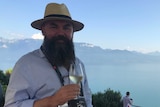 A man in a hat holds up a glass of white wine with a lake in the background