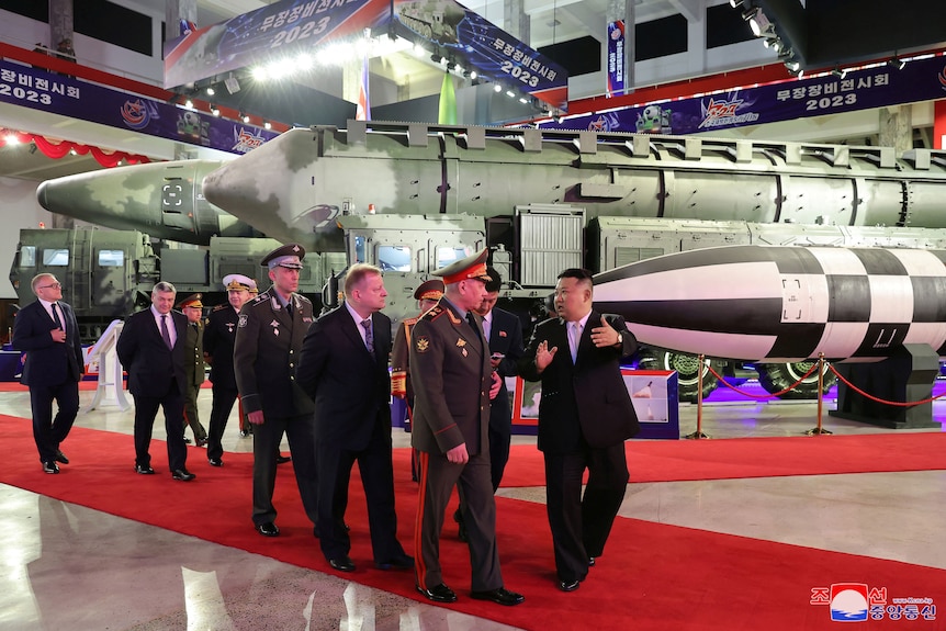 Kim Jong Un leads a group of people including Sergei Shoigu along a red carpet pointing to missiles.