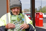 A man with a beanie, holding The Big Issue Magazine, in front of a post office.