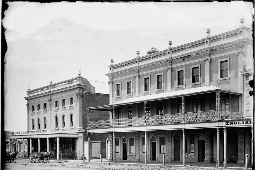 Black and white photograph of two 1800s buildings with a couple of horses and carts in the dirt street in front