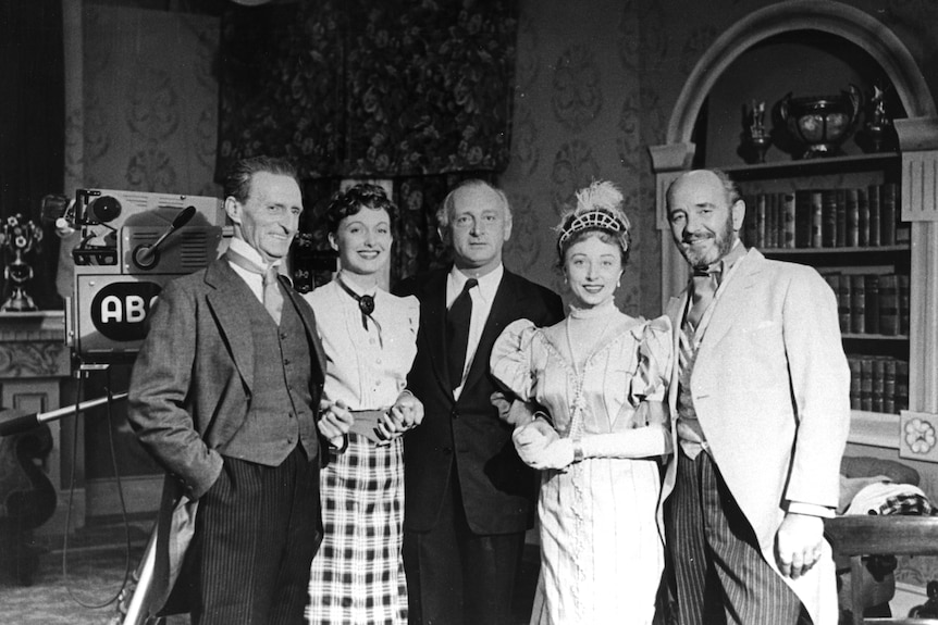 Black and white photo of three men and two women dressed very formally and standing in a line, smiling, with TV camera nearby.