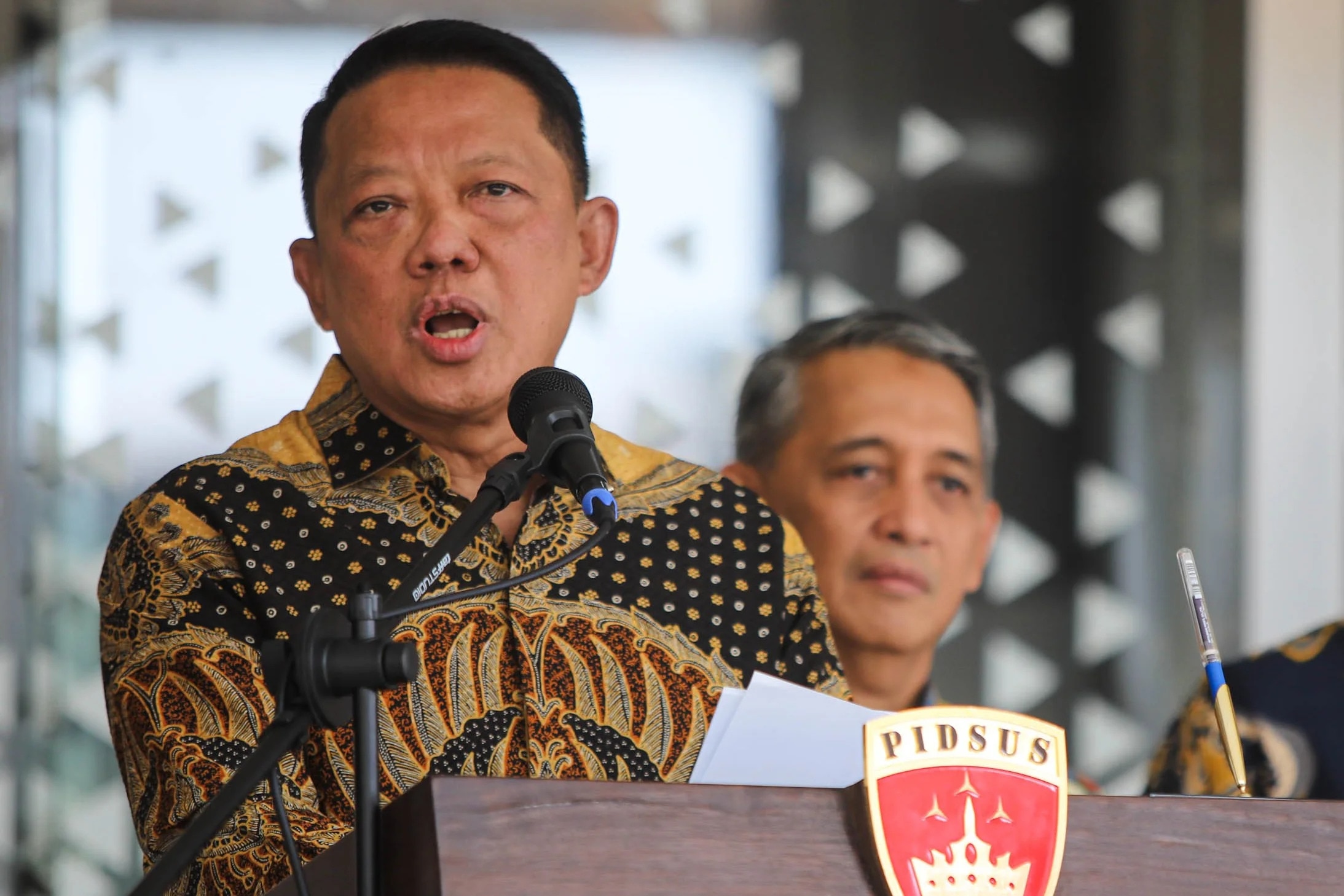 Man wearing a patterned shirt speaking into a microphone, with another man behind him.