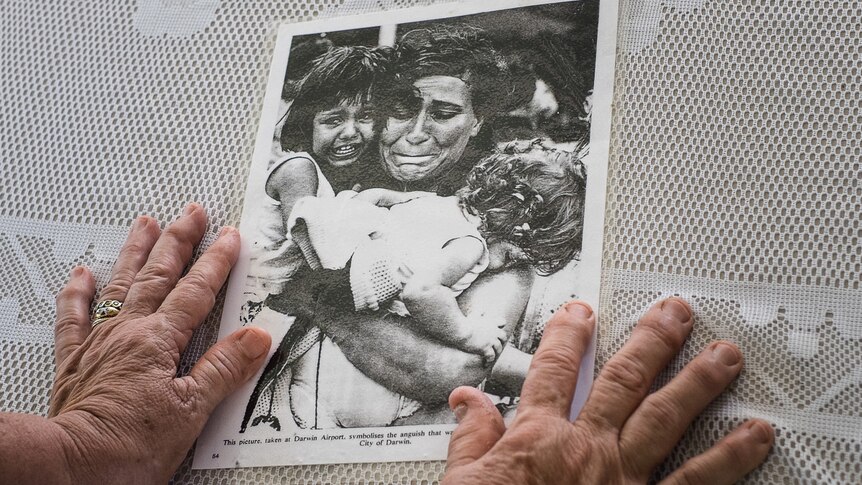 An older woman's hands sit on top of a laminated photograph of a crying woman and children.