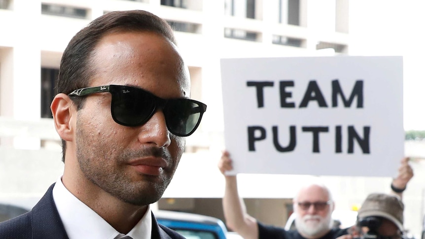 A man in sunglasses is photographed with a protester in the background holding sign that reads "Team Putin"