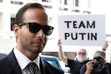 A man in sunglasses is photographed with a protester in the background holding sign that reads "Team Putin"