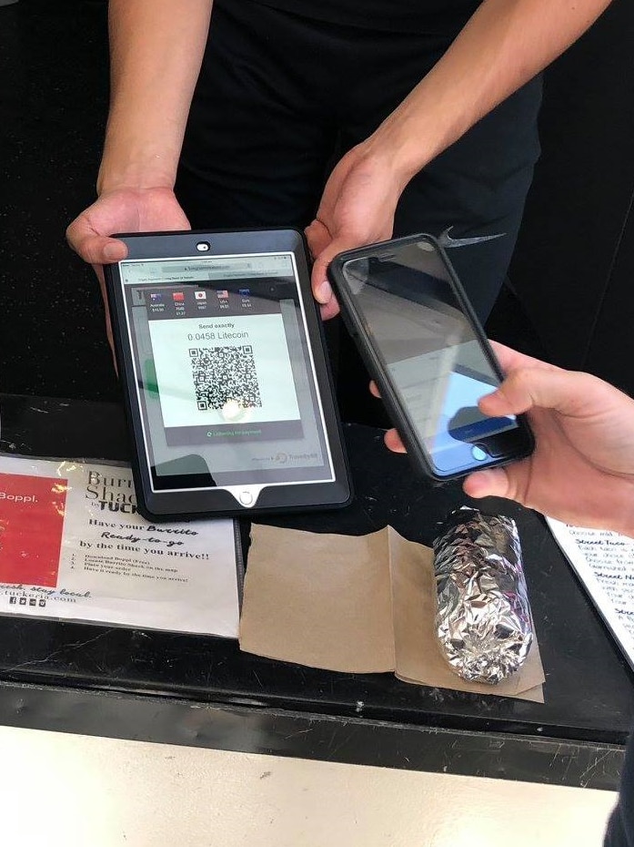 A shop assistant stands behind a counter holding a tablet as a customer holds a smart phone up to it