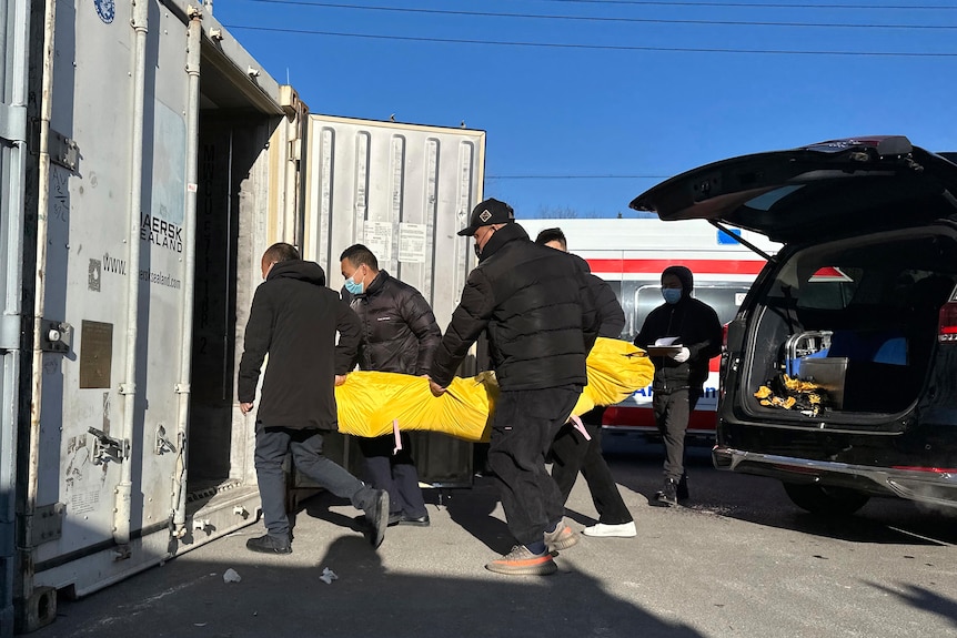 A group of men in winter clothing and respiratory masks carry a body in a bright yellow wrapping into a shipping container.