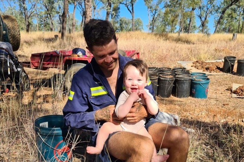 Todd Small holds his baby daughter on his lap, barrels and two vehicles are partially visible in scrub land in the background.
