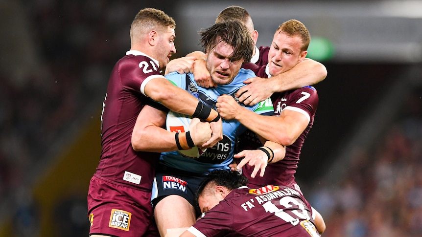 A male New South Wales State of Origin player holds the ball while being tackled by four Queensland opponents.