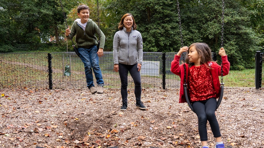 Young mum stands in a park beside her son while pushing her daughter on a swing 