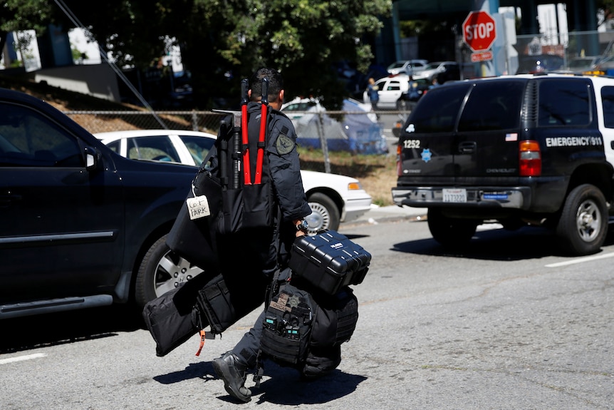 A police officer wearing tactical gear and carrying bags of equipment walks down a street. 