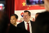 Two men stand smiling for photos before a white backdrop with logos, one is Mark McGowan, people use phone camera.