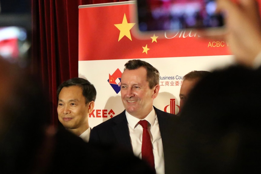 Two men stand smiling for photos before a white backdrop with logos, one is Mark McGowan, people use phone camera.