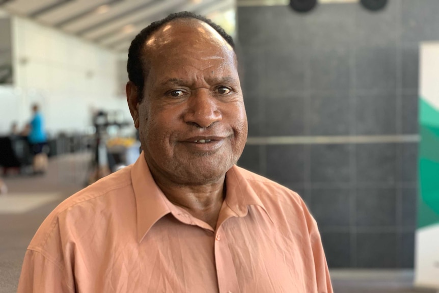 A man with dark hair looks at the camera. He is wearing a peach-coloured shirt