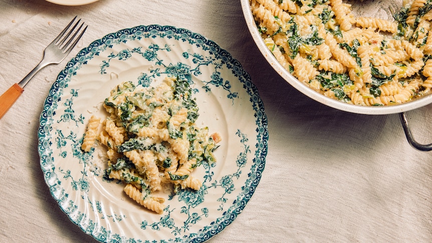 A bowl of creamy green pasta with a fork, next to a larger serving dish.