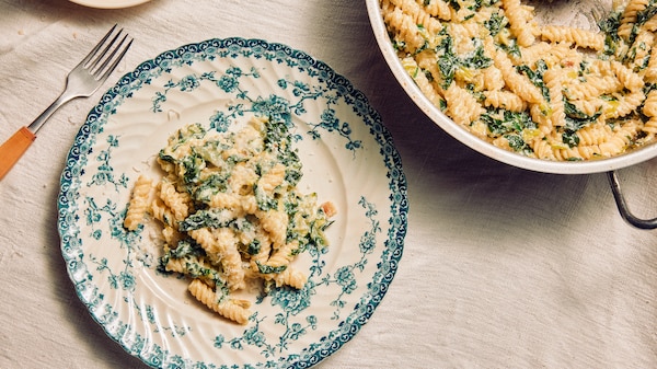 A bowl of creamy green pasta with a fork, next to a larger serving dish.