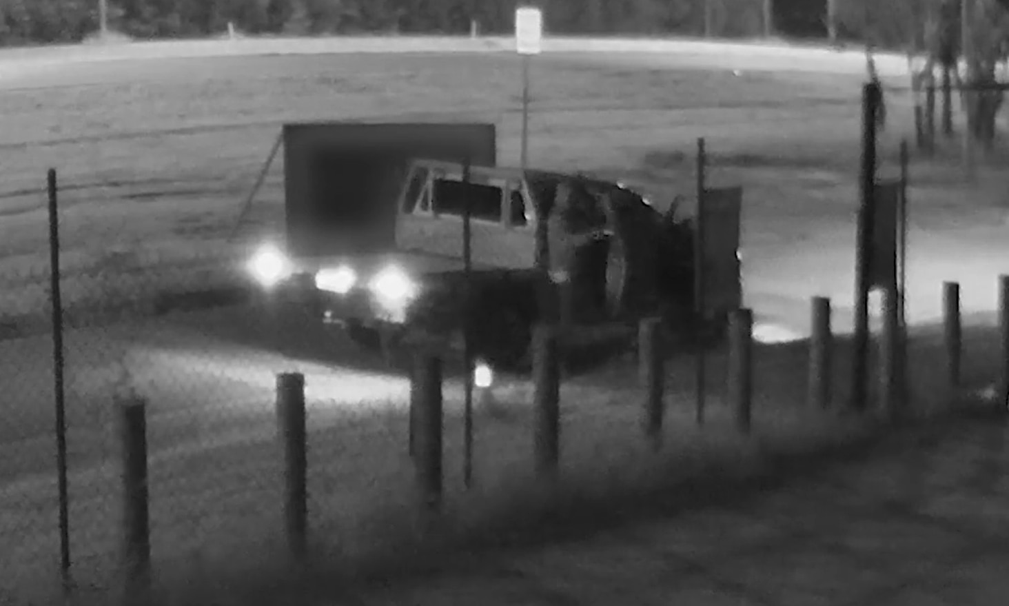 Black and white CCTV of a hooded figure standing next to a ute pointing a gun.