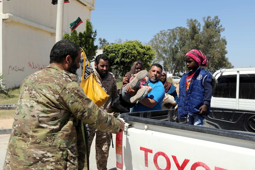 Libyan soldiers assist small children and adults into the back of a white ute on a bright blue day in Tripoli's conflict areas.