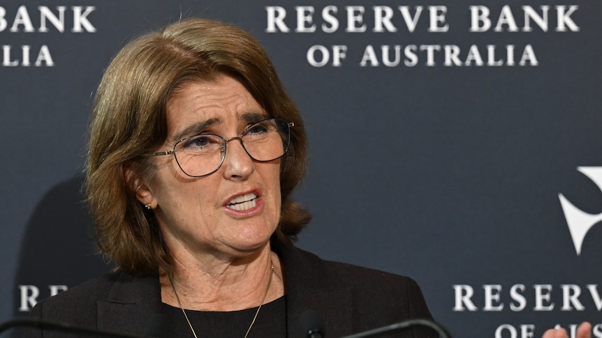 A woman with short brown hair and glasses speaks in front of a media wall that says "Reserve Bank of Australia".