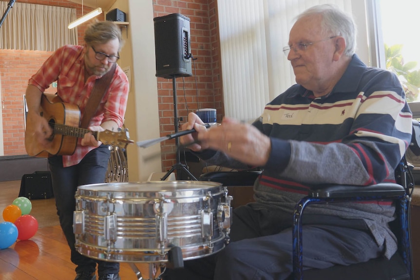 An eldrly drummer performing with a much younger guitarist in an indoor setting