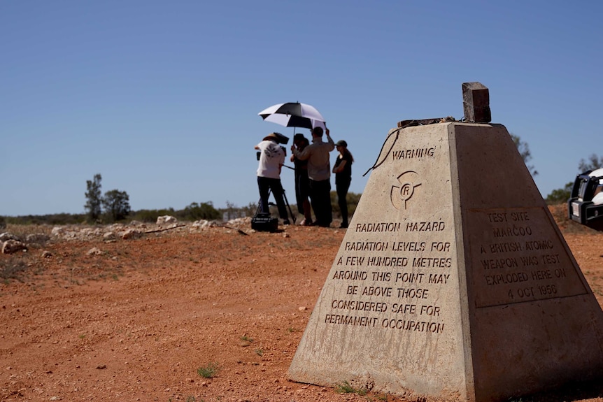 A film crew huddle an umbrella near a concrete block marking the Maralinga nuclear test site.