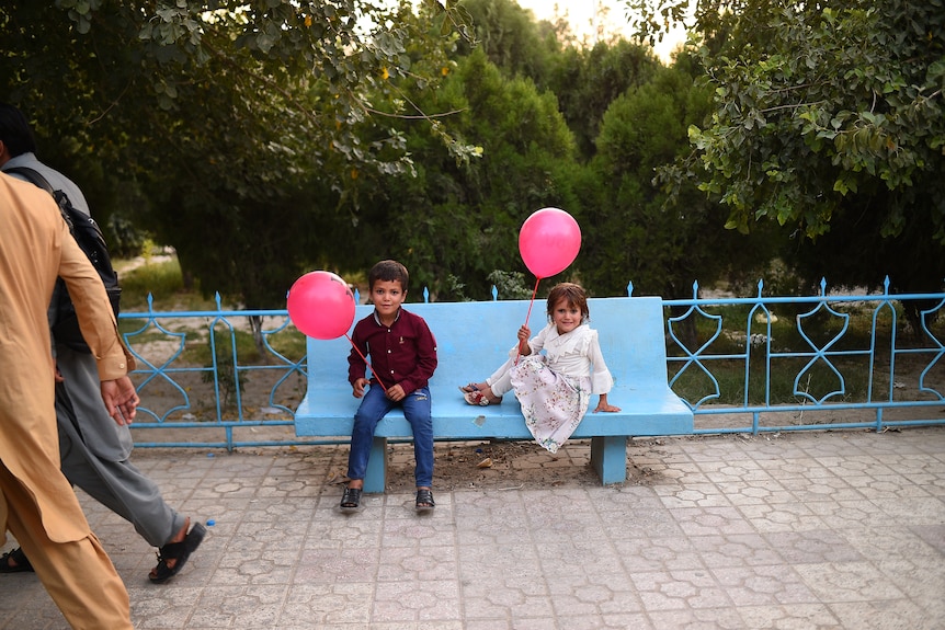 A young boy and girl sit on a park bench holding bright pink balloons. 