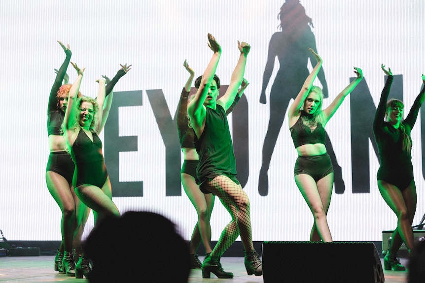 Colour photograph of Bey Dance performers in black outfits dancing on stage in front of an LED screen.