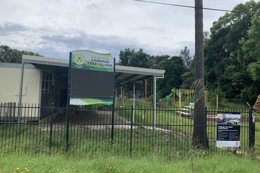the front of Cabbage Tree Island School is shown with a sign and school buildings behind a gate