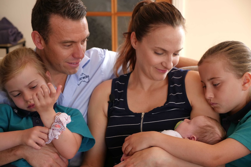 A mother and father sit on a lounge with their two young daughters, and baby.