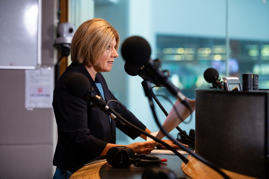 A woman with short blonde hair with a black jacket and light blue shirt stands in front of microphones speaking.
