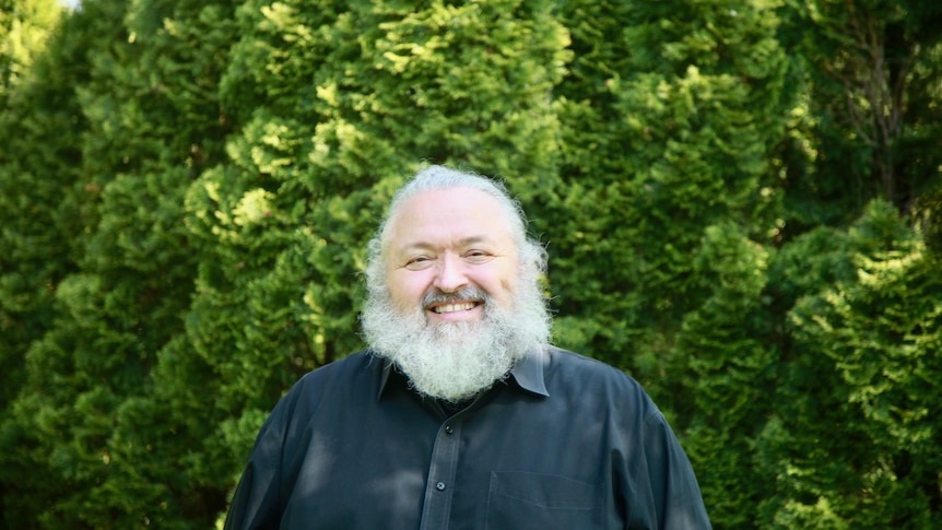 You view a man with white hair and a thick, long beard smiling in front of a verdant hedge.