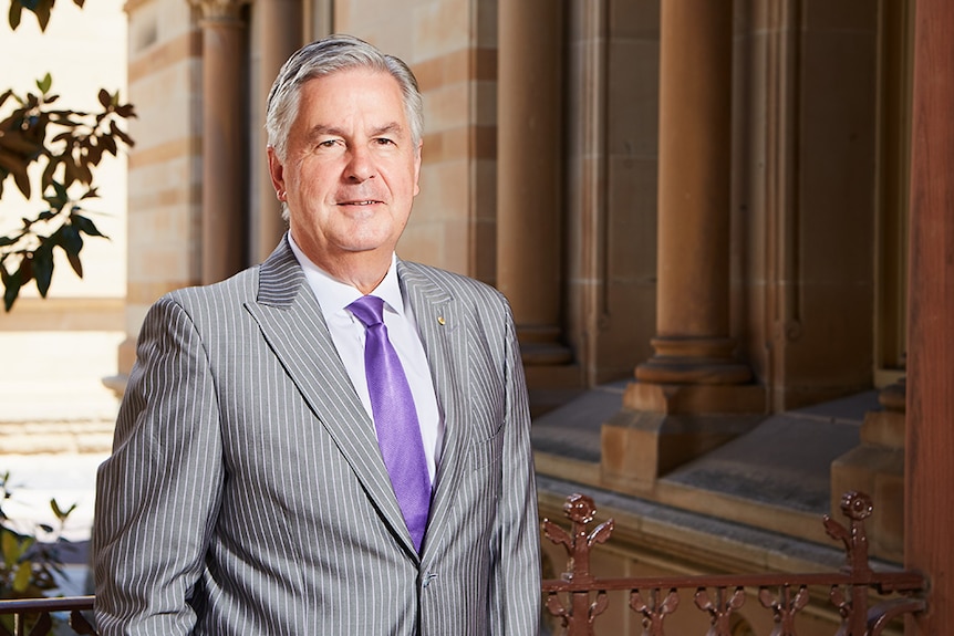 A man with grey hair and a suit, with a purple tie, stands in front of a building with sandstone columns.