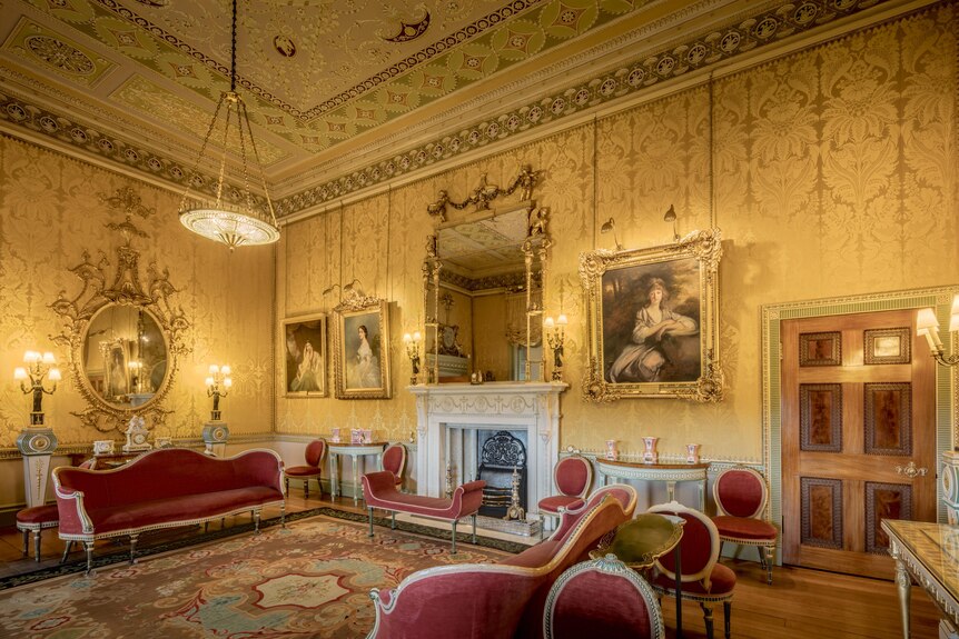 A very opulent old-fashioned room, with a fireplace, gold wallpaper, art and mirrors in gold frames and red lounges