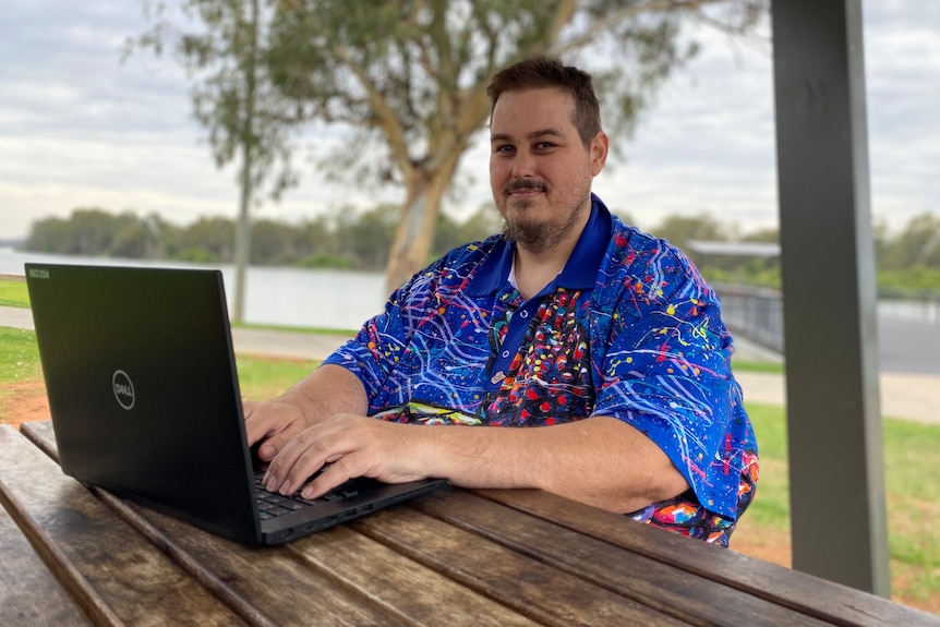 A man has short brown hair and wears a colourful tshirt, he sits at a wooden table in a park and types at a black computer