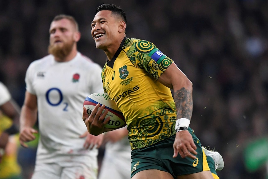 A man in a gold jersey smiles as he runs along with a rugby ball under his arm