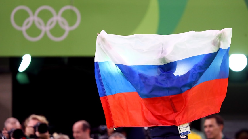 An athlete is obscured by a Russian flag they are holding up at the Olympic Games. The Olympic rings are visible in the top left
