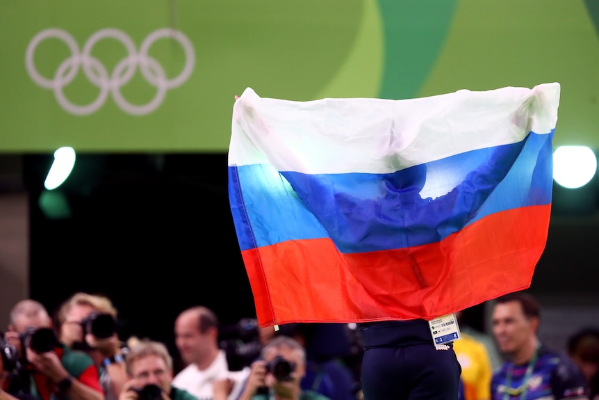 An athlete is obscured by a Russian flag they are holding up at the Olympic Games. The Olympic rings are visible in the top left