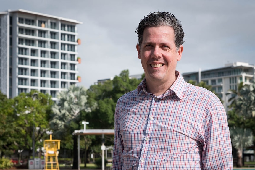 A man in a checked shirt smiles in front of the Cairns Lagoon with apartments behind.