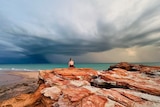 A woman sits on red rocks over a dark ocean and stormy sky