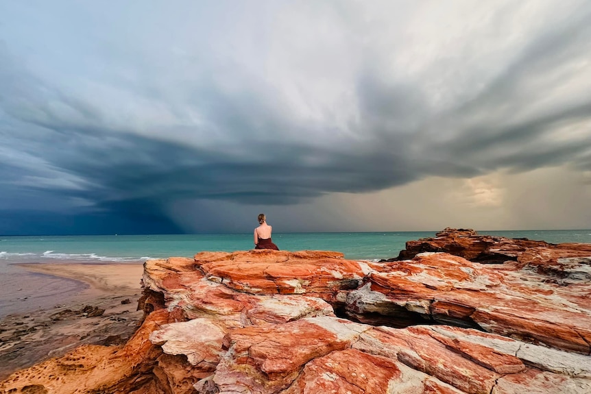 A woman sits on red rocks over a dark ocean and stormy sky