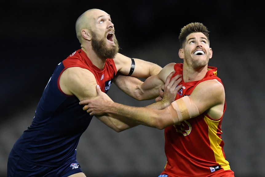 A bald AFL players in blue and red wrestles with a shorter player in orange as they look into the sky.